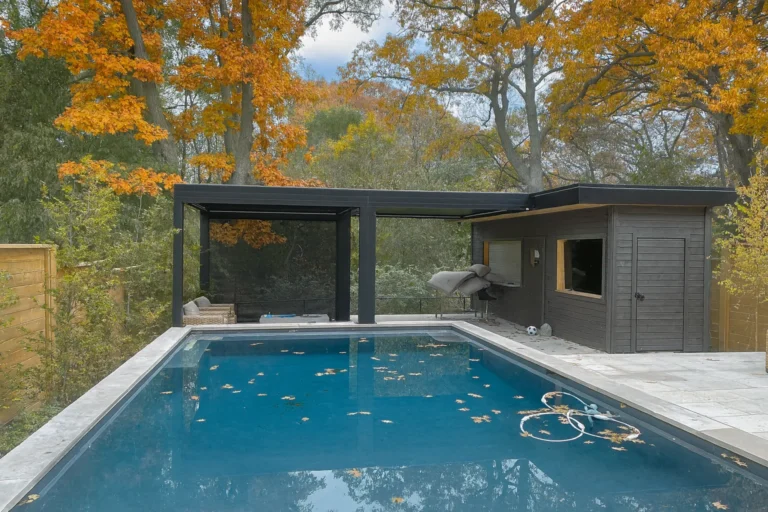 Black louvered pergola over a poolside lounge area beside a pool house, surrounded by autumn foliage
