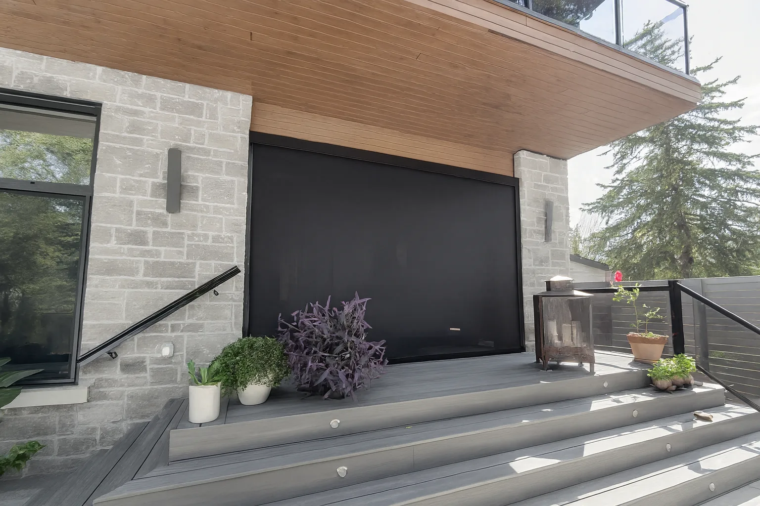 Large black retractable screen fully lowered across a stone and wood-clad house patio with potted plants