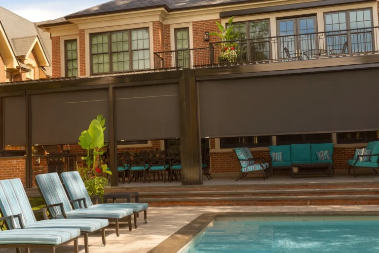 Dark retractable mesh screens lowered along a louvered pergola patio beside a pool with striped chaise lounges