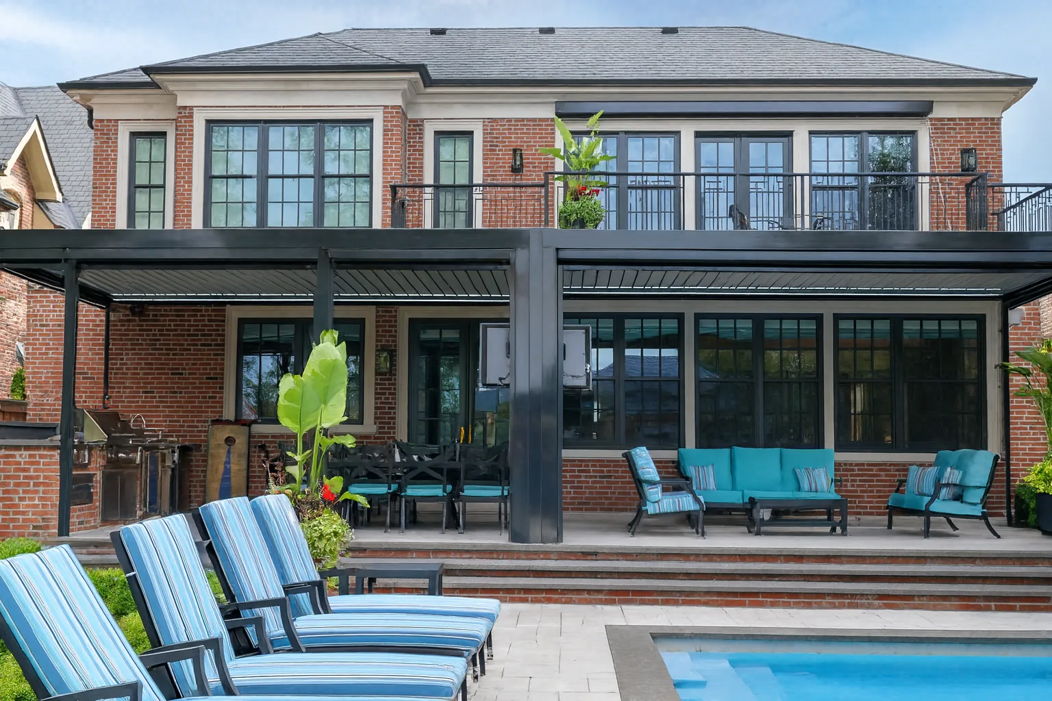 Two large dark-framed louvered pergolas spanning the rear patio of a brick home beside a pool